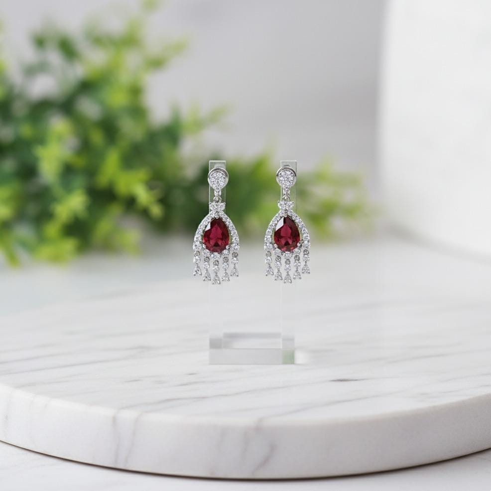 Pair of earrings with red gemstones on a white surface with greenery in the background