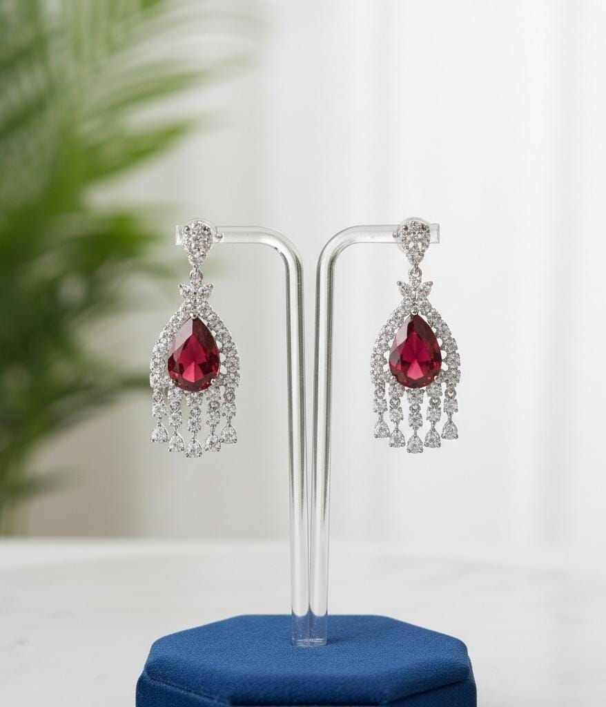 Pair of red teardrop earrings on a clear stand with a blurred green plant in the background.