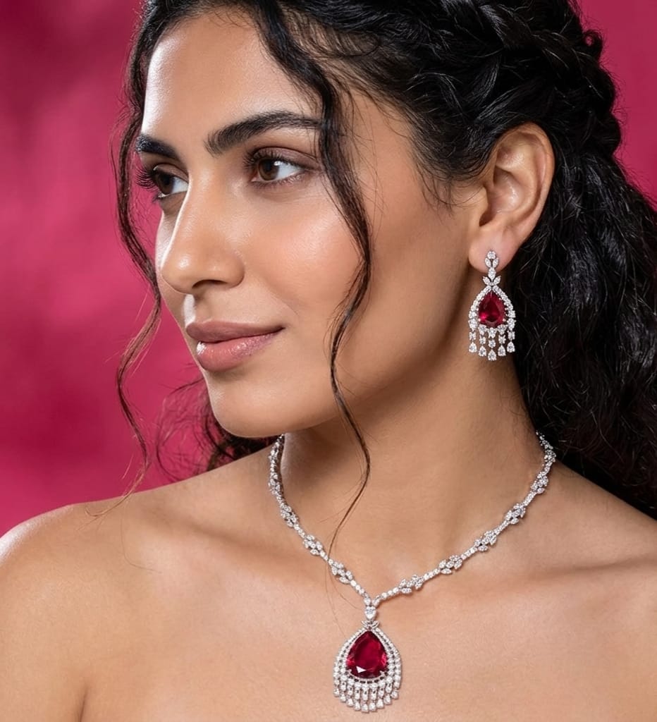 A woman wearing a matching jewelry set featuring a teardrop-shaped red gemstone pendant necklace and matching drop earrings with clear stones, set against a solid pink background.