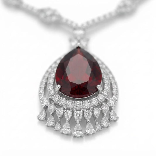 A silver pendant necklace featuring a teardrop-shaped deep red gemstone surrounded by a halo of small clear stones, with dangling teardrop-shaped clear stones below, set against a plain white background.