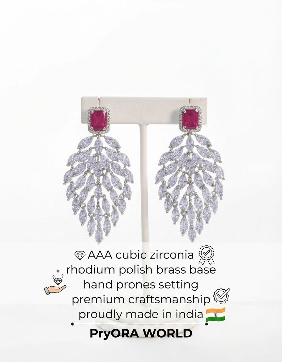 A pair of large drop earrings displayed on a white stand against a white background, with product text below. The earrings have rectangular pink gemstones at the top and cascading leaf-shaped clusters of clear stones on a silver-toned branch.